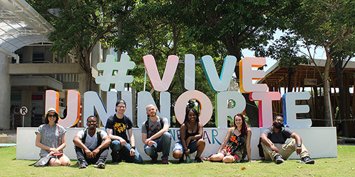 Seven students posing casually for a photo in front of a sculpture with text in Spanish