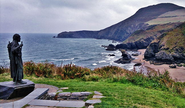 Dramatic scene of a statue overlooking the grass, rocks, and sand of the Welsh coastline.